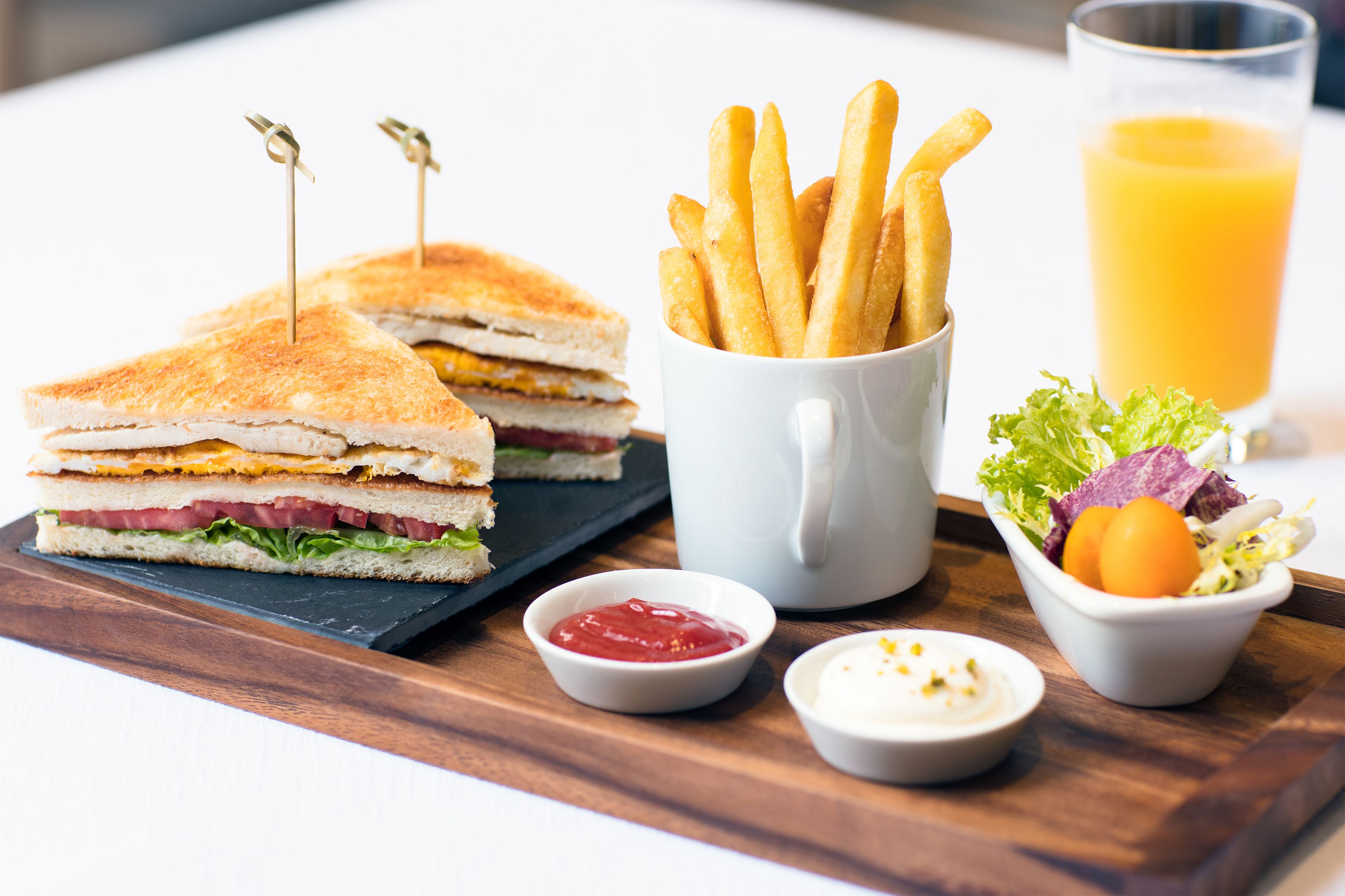 Un repas composé d’un sandwich, de frites, d’un verre de jus d’orange et de petits ramequins de ketchup et de sauce crémeuse.