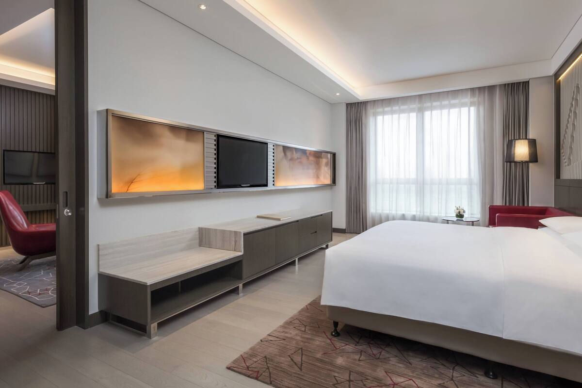 Una habitación de hotel moderna con una cama grande, un televisor de pantalla plana montado en la pared, una zona de estar con un banco y una mesita, y un ventanal con cortinas.