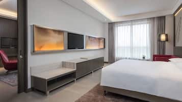 A modern hotel room with a large bed, a flat-screen TV mounted on the wall, a seating area with a bench and a small table, and a large window with curtains.
