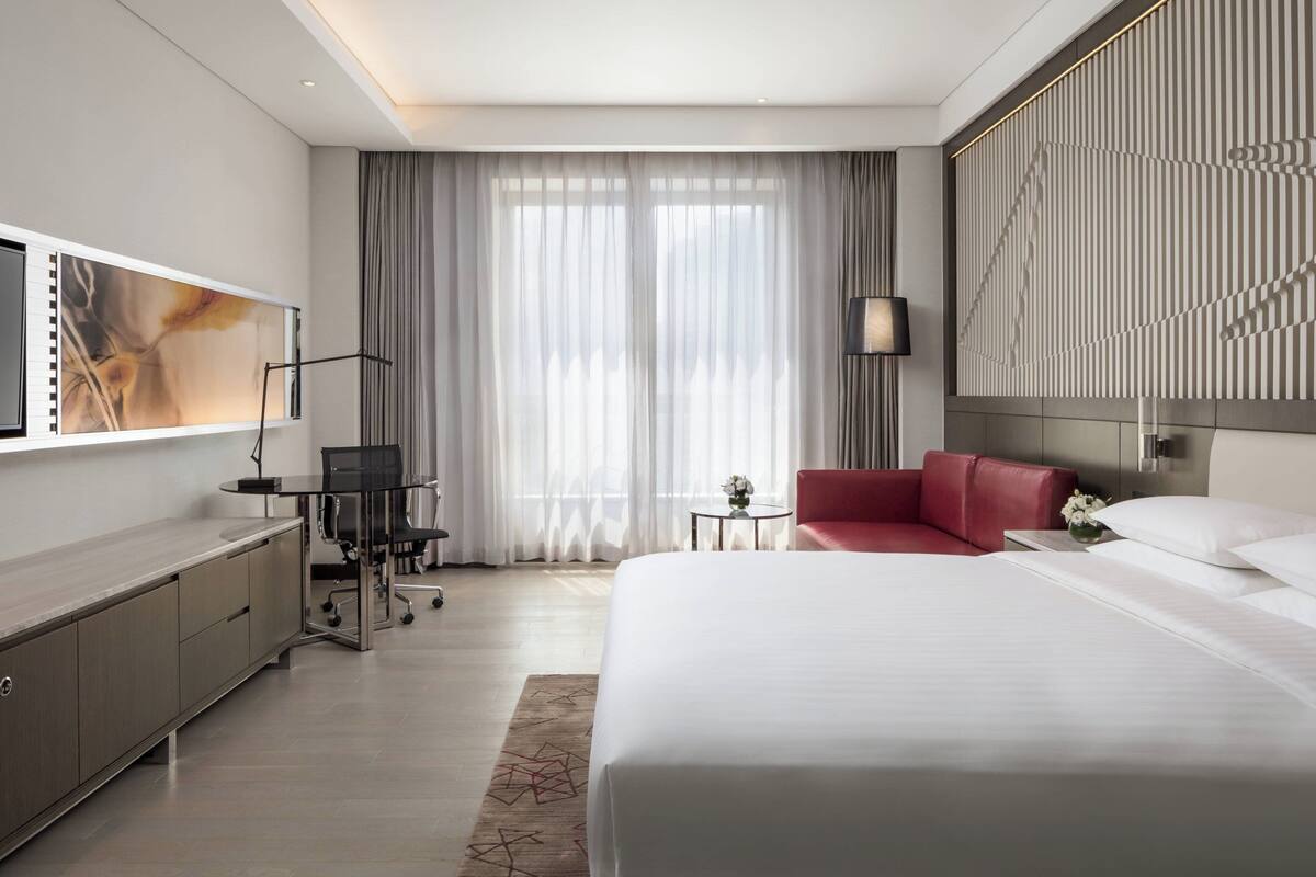 Una habitación de hotel moderna con una cama grande, un escritorio con una silla, un sofá rojo, un televisor montado en la pared y un ventanal con cortinas.