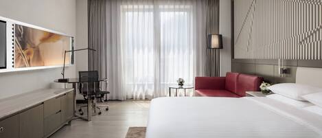 A modern hotel room with a large bed, a desk with a chair, a red sofa, a TV mounted on the wall, and a large window with curtains.