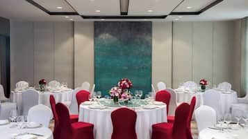 A banquet hall with round tables set for dining, white and red chairs, and a large abstract painting on the wall.