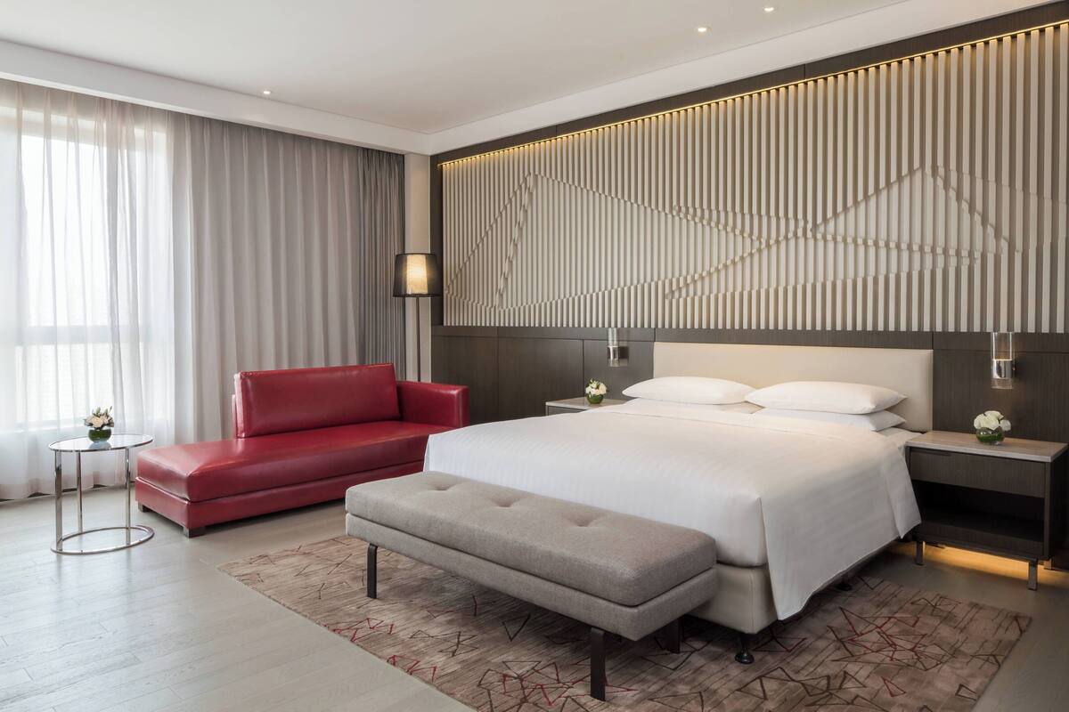 Una habitación de hotel moderna con una cama grande, un sofá rojo, una mesa redonda pequeña y un banco.
