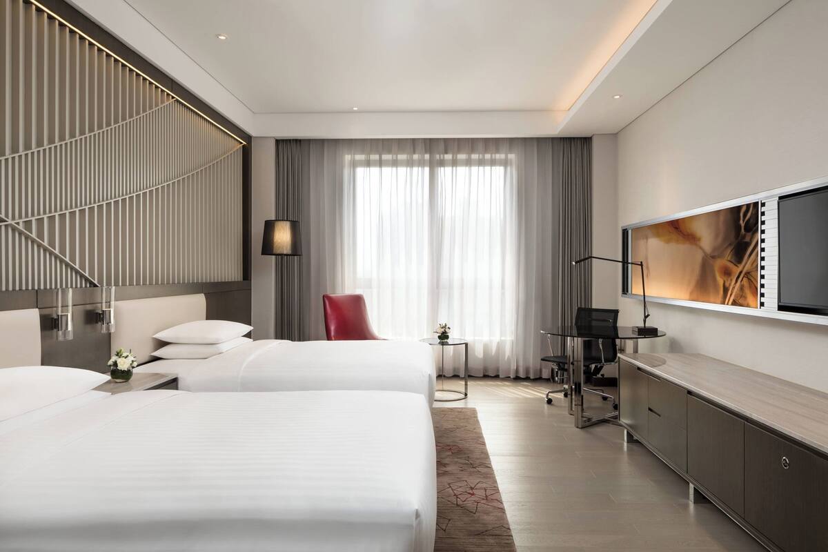 Una habitación de hotel moderna con dos camas, un escritorio, un televisor y un ventanal grande con cortinas.