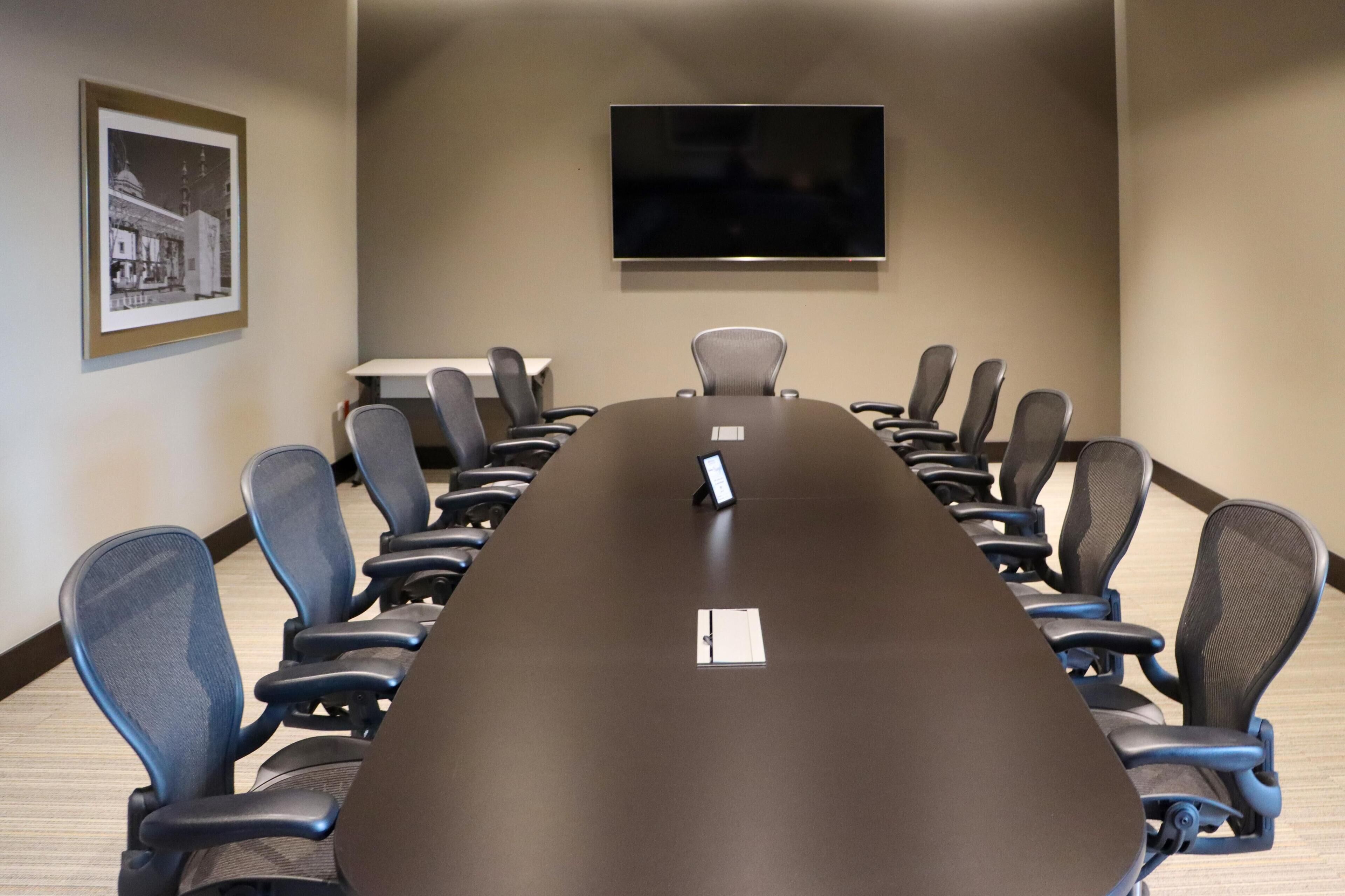 A conference room with a long table, chairs, a mounted TV, and a framed picture on the wall.