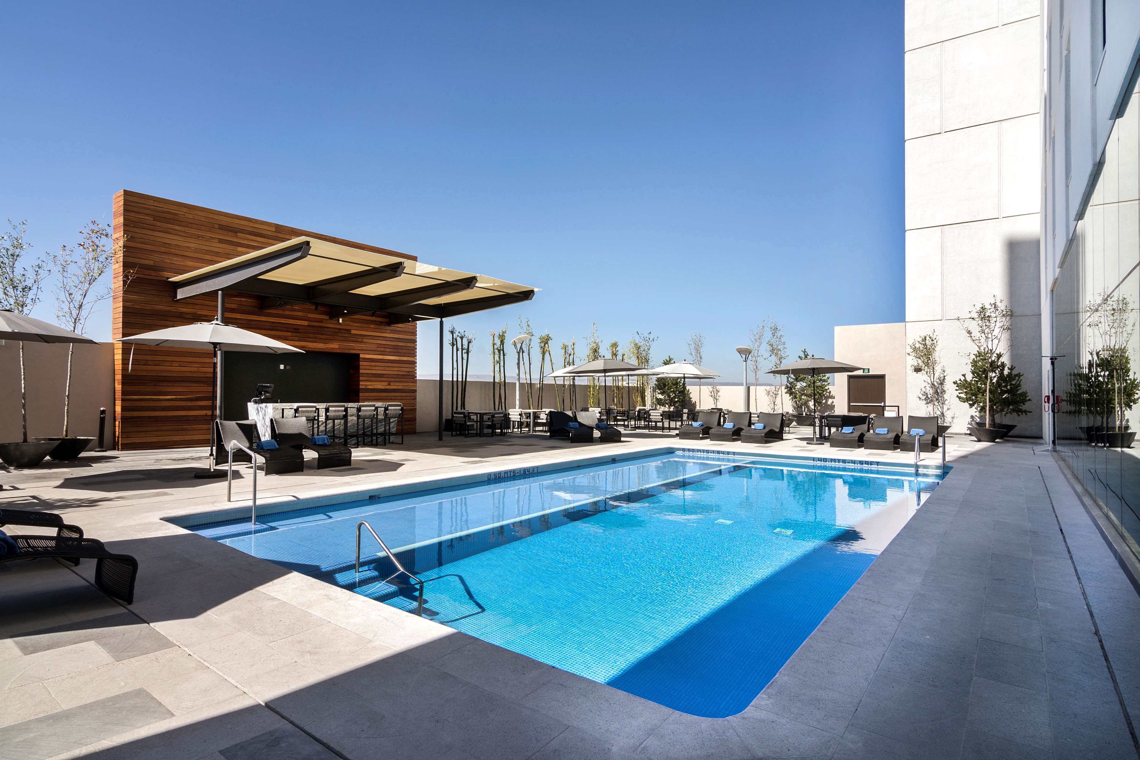 A modern outdoor pool area with lounge chairs and umbrellas, surrounded by a building with a wooden facade and a clear blue sky.