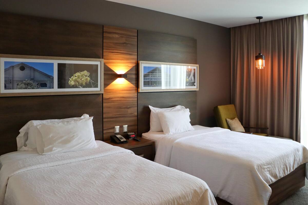 Habitación de hotel con dos camas, una mesita de noche con teléfono y lámpara, y una obra de arte colgada en la pared.