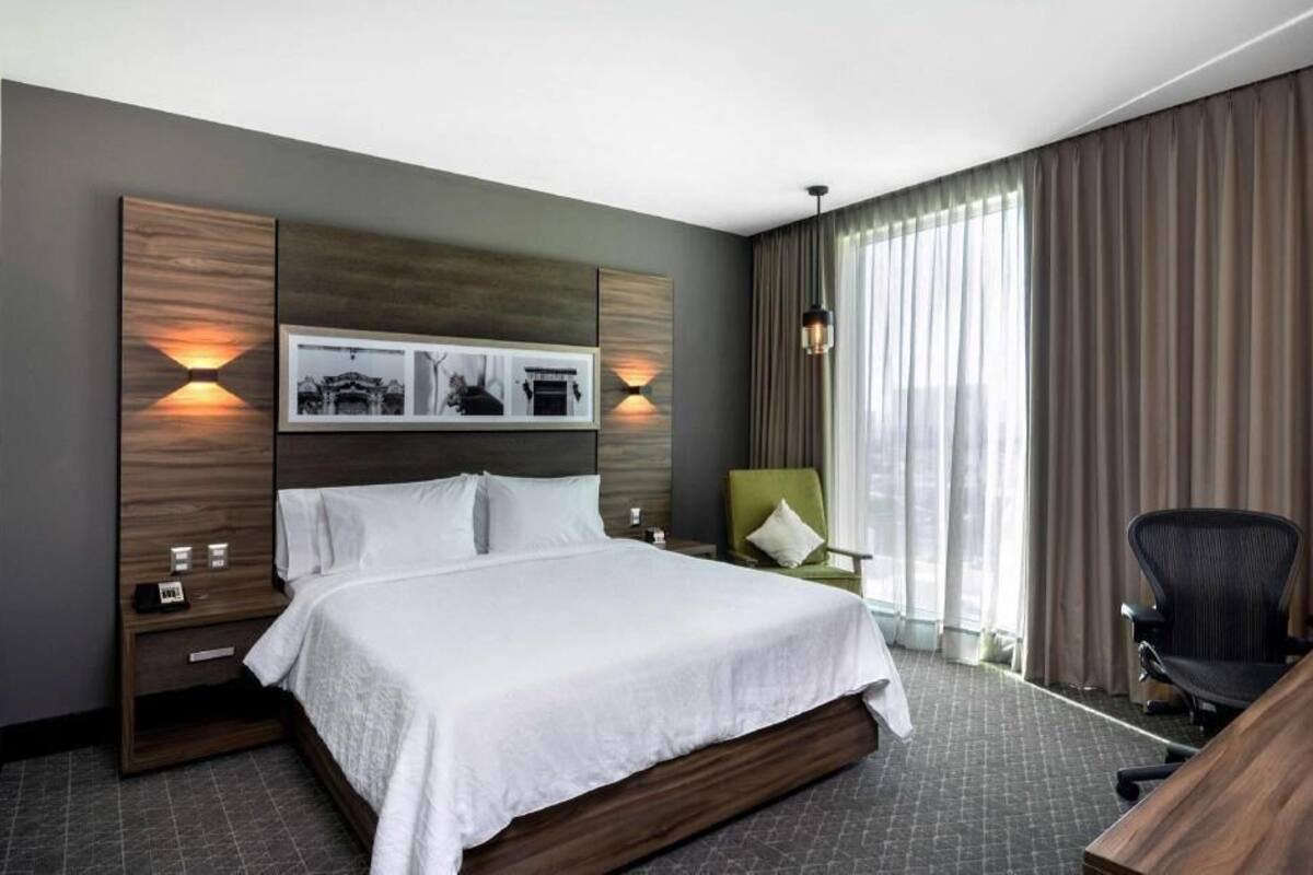 Una habitación de hotel moderna con una cama grande, un escritorio, una silla y vistas de la ciudad a través de la ventana.