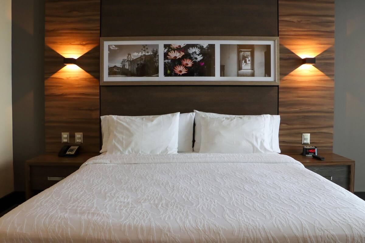 Una cama bien hecha con sábanas blancas, un teléfono en la mesita de noche y un reloj digital sobre un mueble de madera.