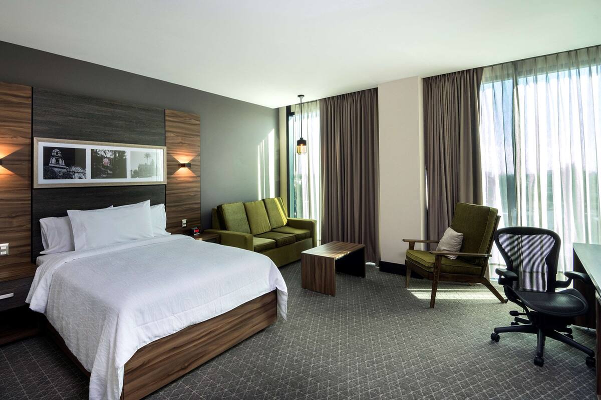 Una habitación de hotel con una cama grande, un sillón verde, una mesa de centro de madera y una silla de oficina negra.
