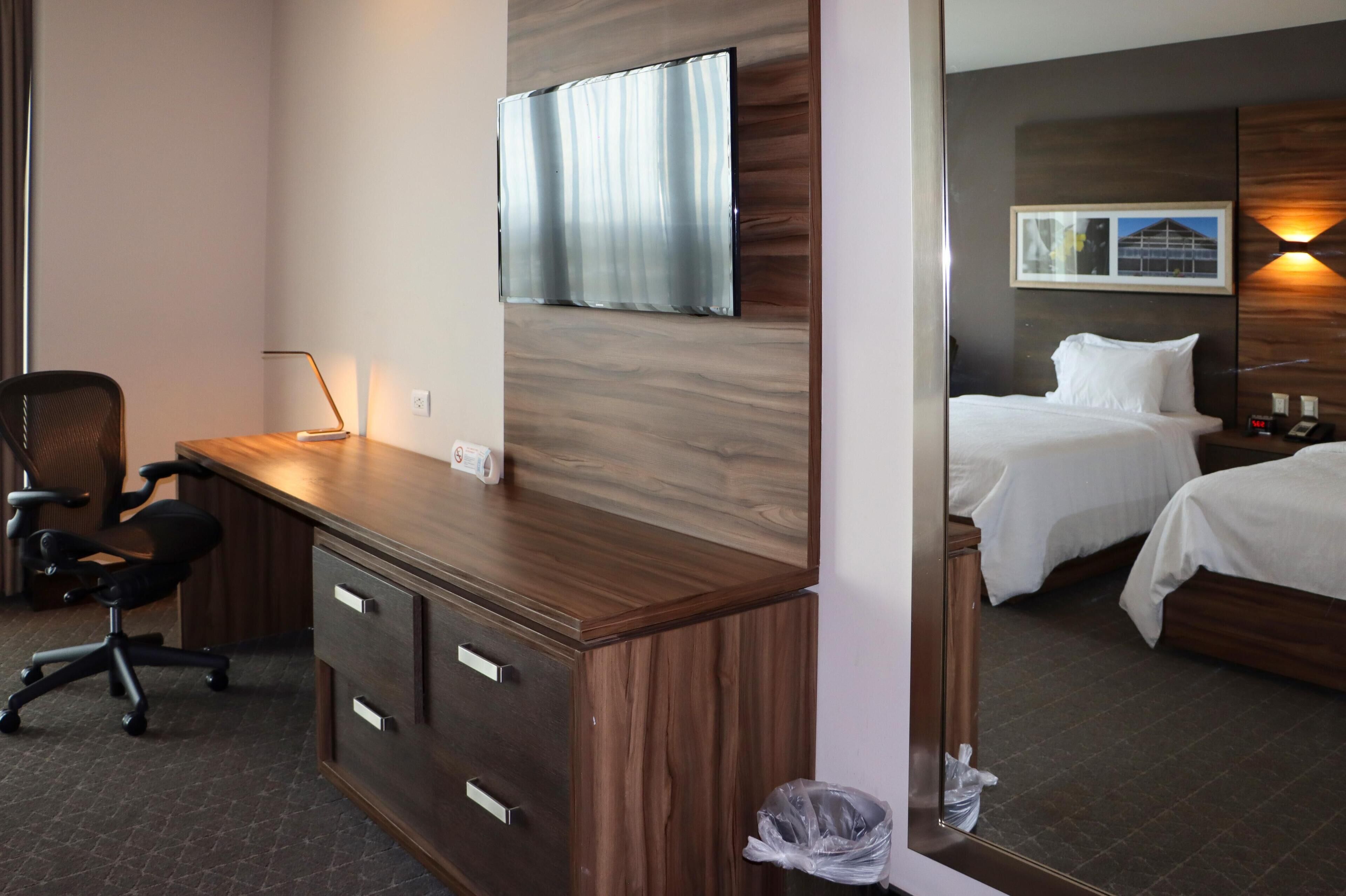 A hotel room with a wooden desk, two beds, a TV, and a framed picture on the wall.