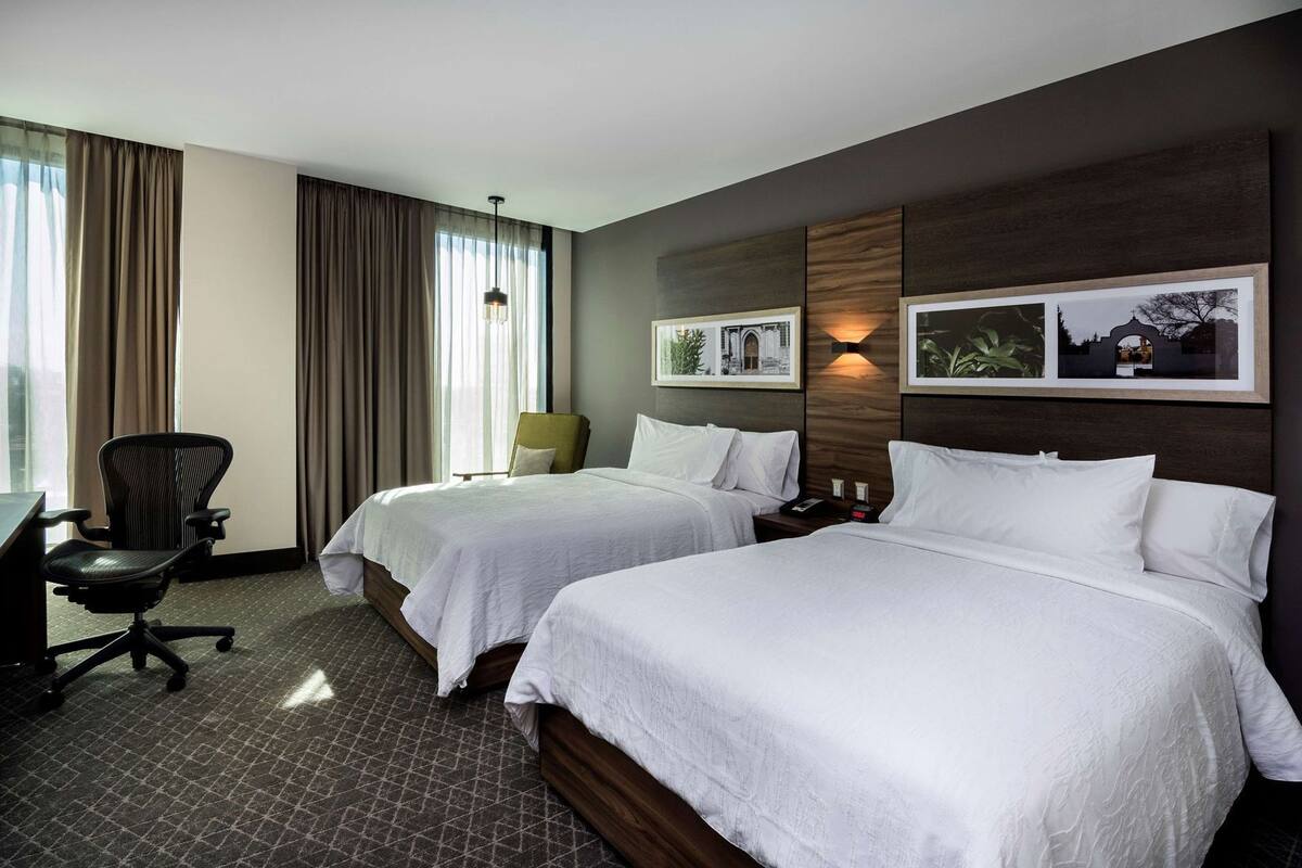 Habitación de hotel con dos camas, un escritorio, una silla y un ventanal con cortinas.