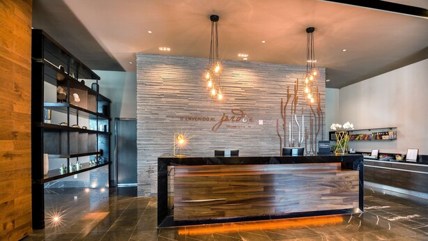 A modern hotel reception area with a wooden reception desk, a wall-mounted light fixture, and a shelving unit with various items. - Hilton Garden Inn Aguascalientes (Aguascalientes)