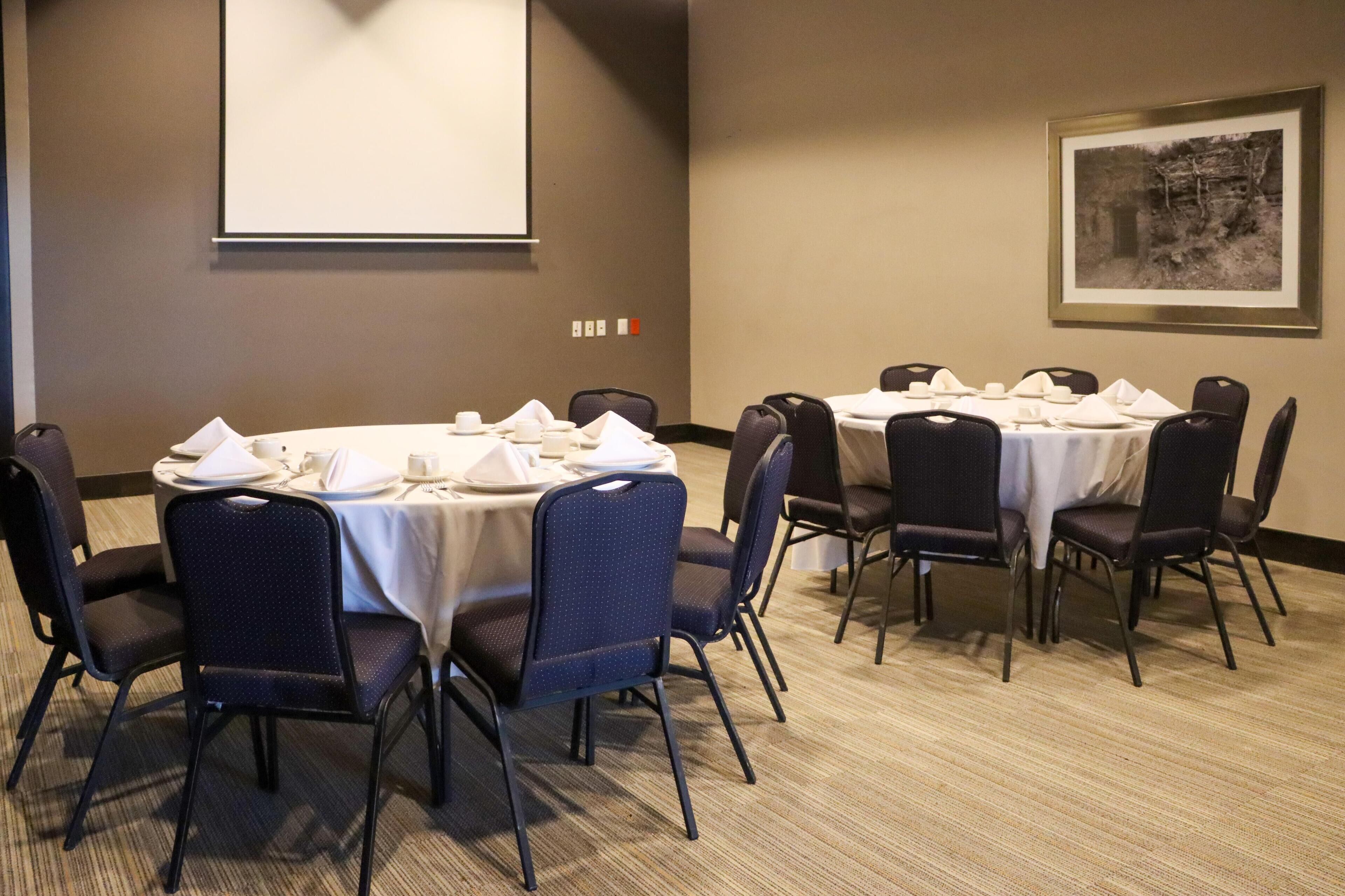 A conference room with round tables, chairs, a projector screen, and a framed picture on the wall.
