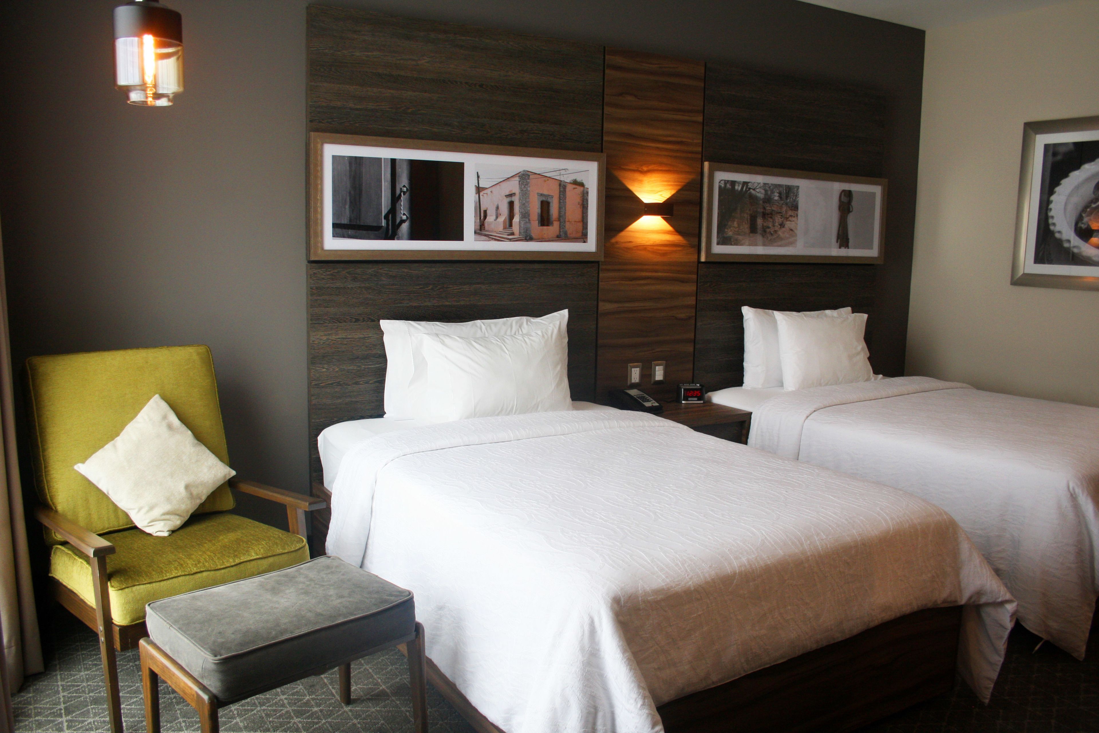 A hotel room with two beds, a green armchair, and a small table.