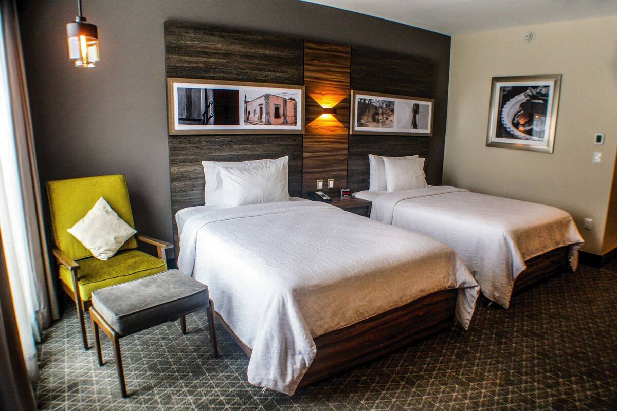 Una habitación de hotel con dos camas, un sillón verde, una mesita y cuadros colgados en la pared.