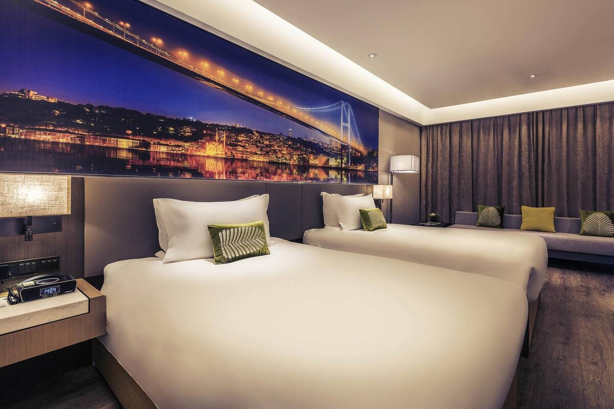 A hotel room with two beds, a nightstand, a lamp, and a large cityscape mural on the wall.