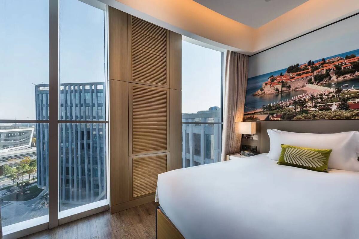 A hotel room with a large window, a bed, a nightstand, a painting, and a view of buildings.