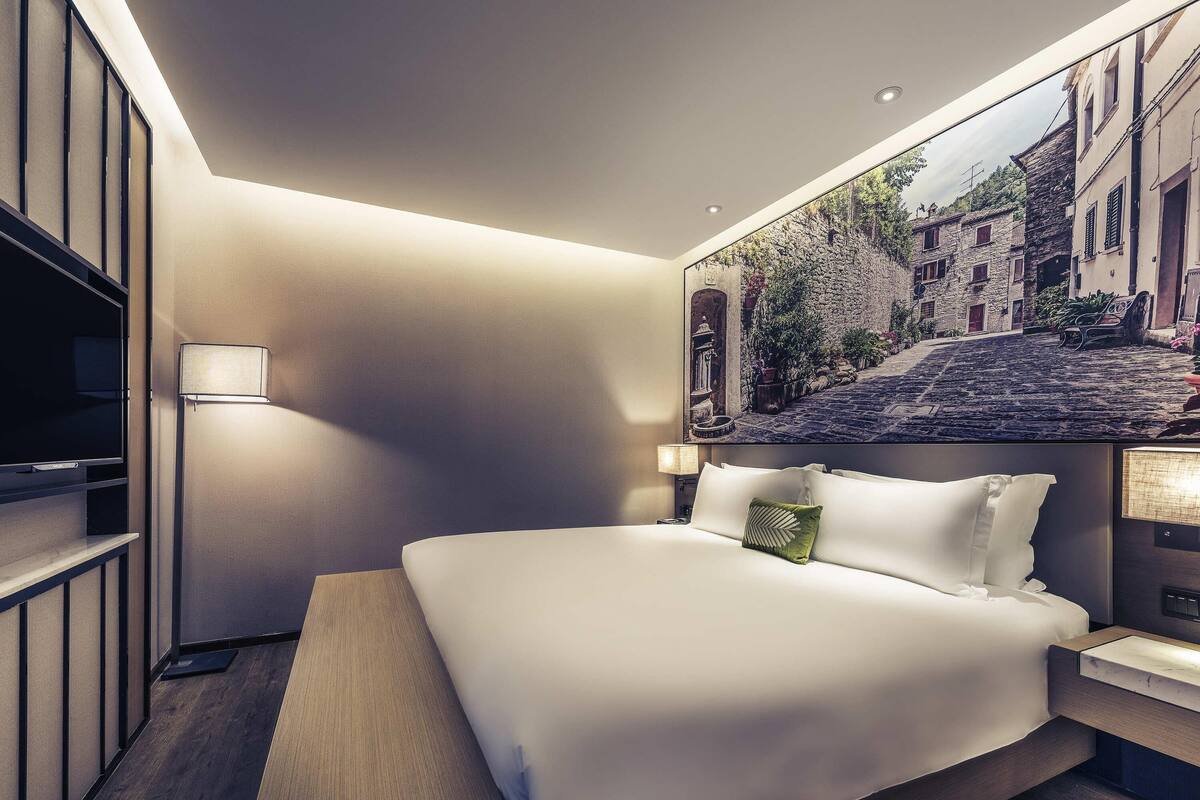 A modern hotel room with a large bed, a flat-screen TV, a painting of a street scene, and a wooden bench.