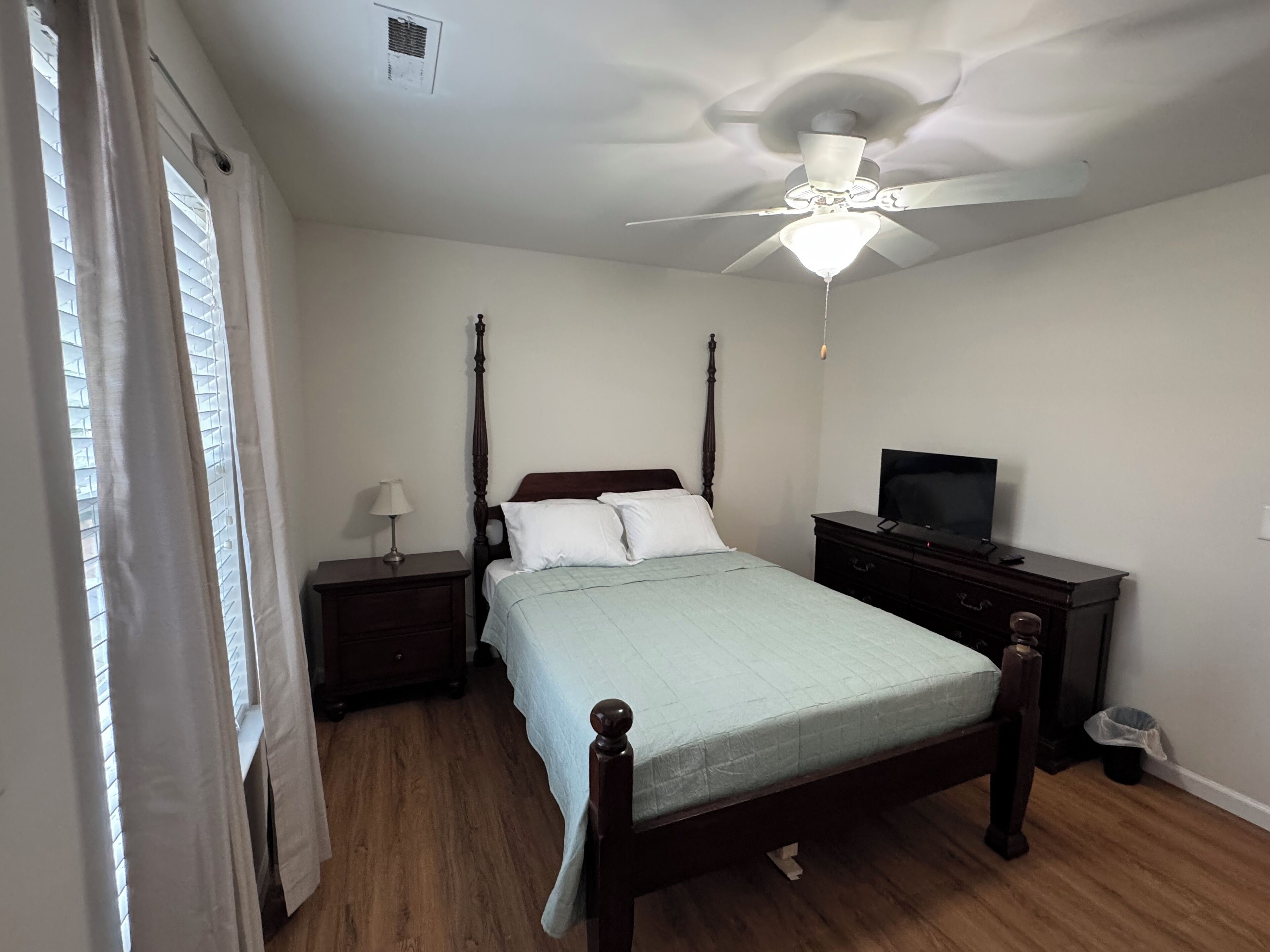 A bedroom with a four-poster bed, a ceiling fan, a nightstand with a lamp, a dresser, and a television.
