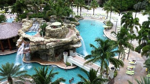 Área de piscina en un complejo turístico con una cascada, palmeras y sillas de descanso.