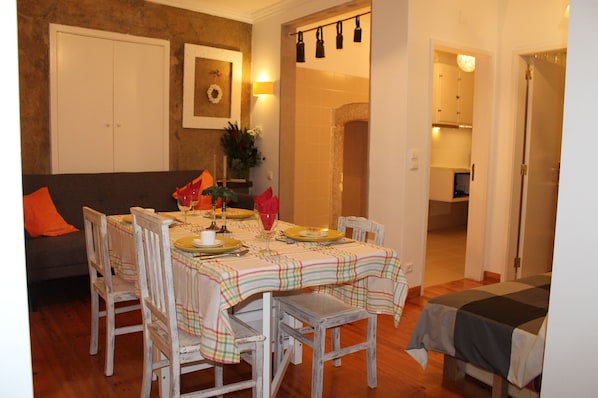 A dining area with a table set for four, surrounded by wooden chairs. A sofa with an orange cushion is visible in the background.
