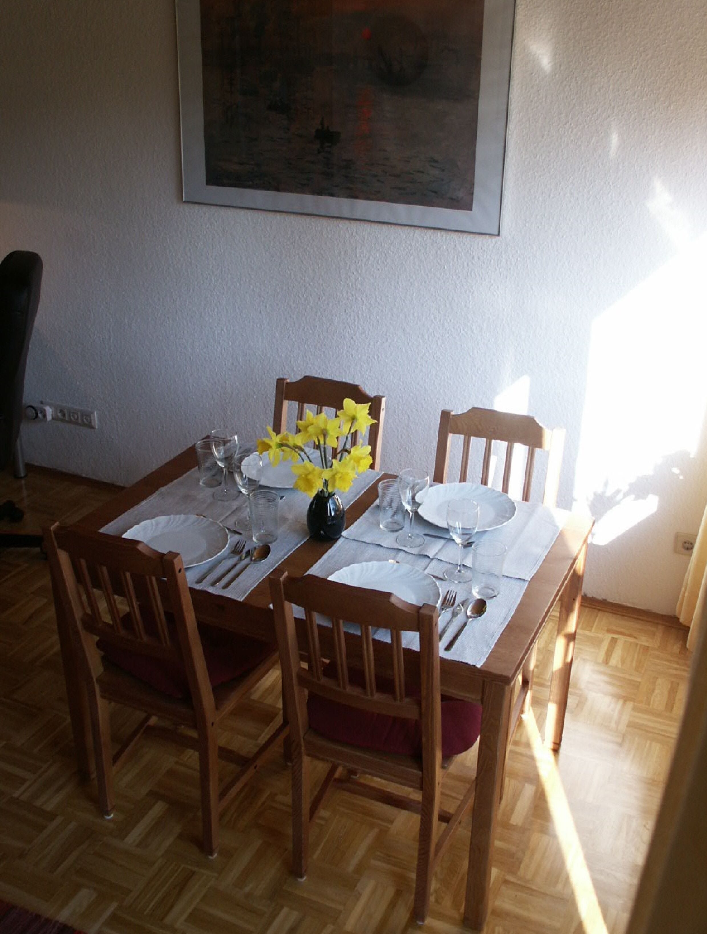 A dining table set for four with chairs, a vase of yellow flowers, and a painting on the wall.