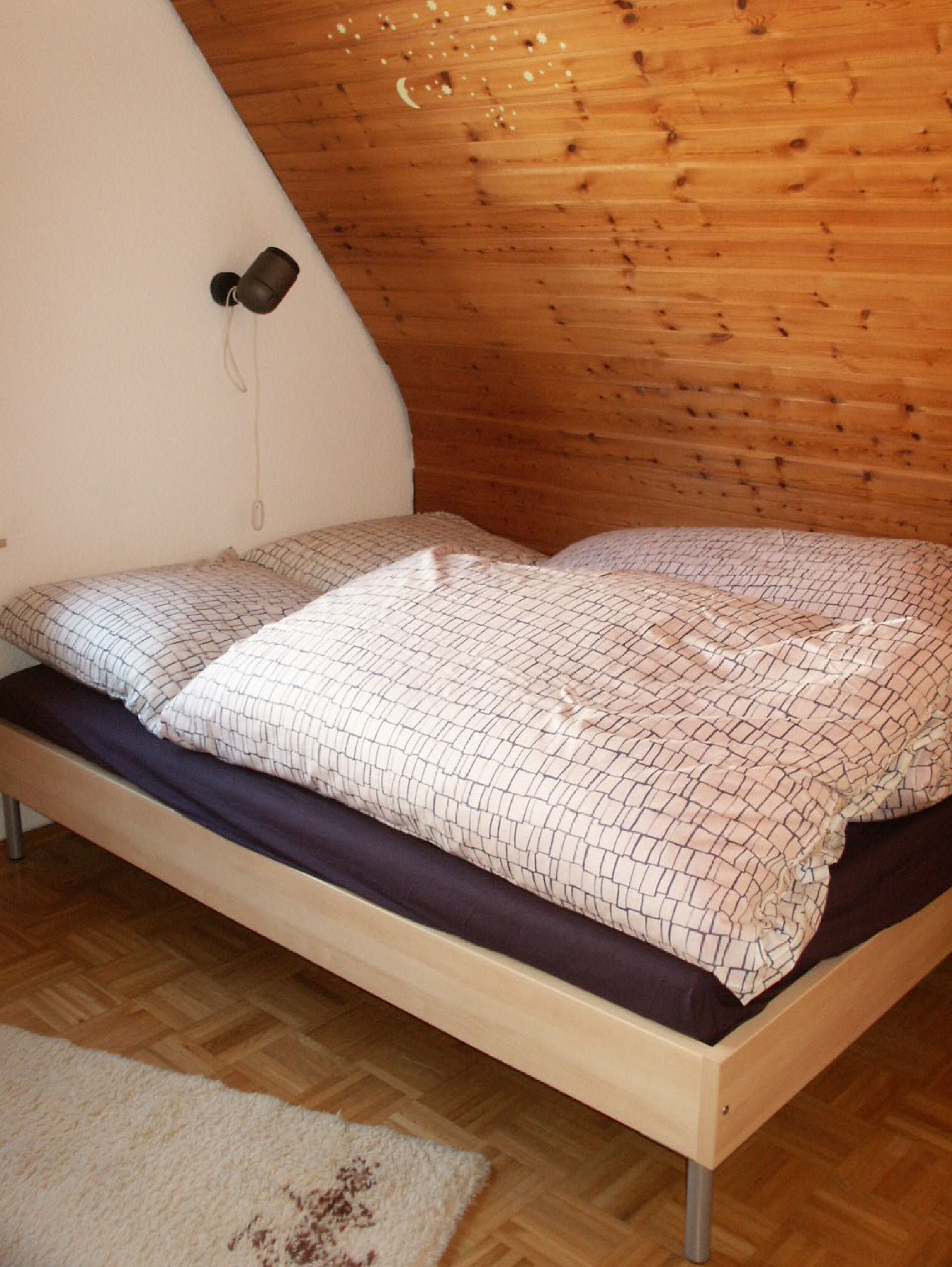 Una cama individual con colcha y almohadas estampadas, suelo de madera y pared blanca con toma de corriente.