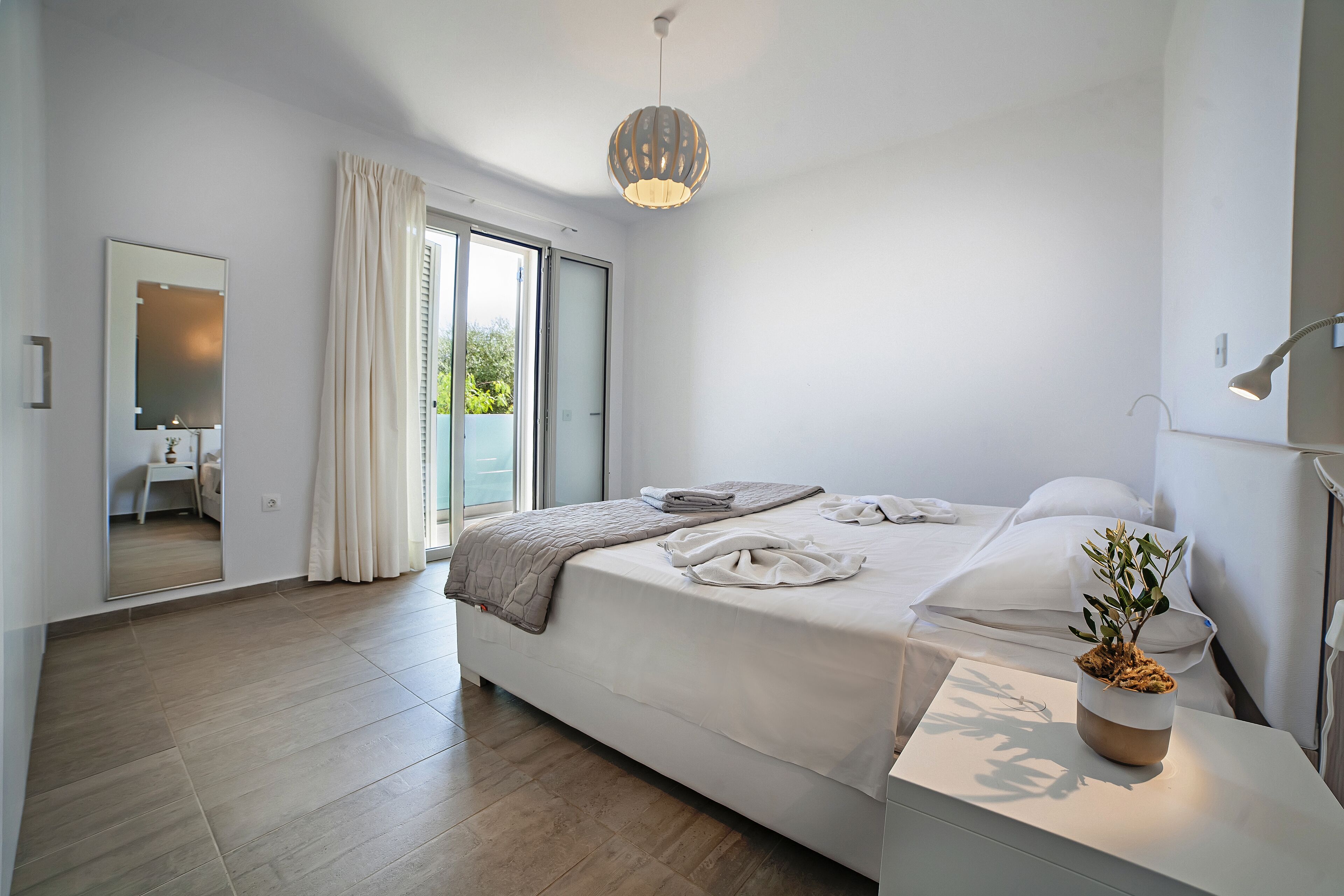 A modern bedroom with a large bed, a bedside table with a plant, and a view of the outdoors through a sliding door.
