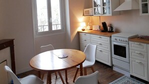 Una cocina compacta con armarios blancos, una mesa de madera con cuatro sillas, un ventanal y una lámpara en el techo.