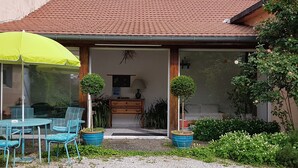 Una casa con techo de tejas, un patio con sillas azules y un paraguas amarillo, y plantas en macetas.