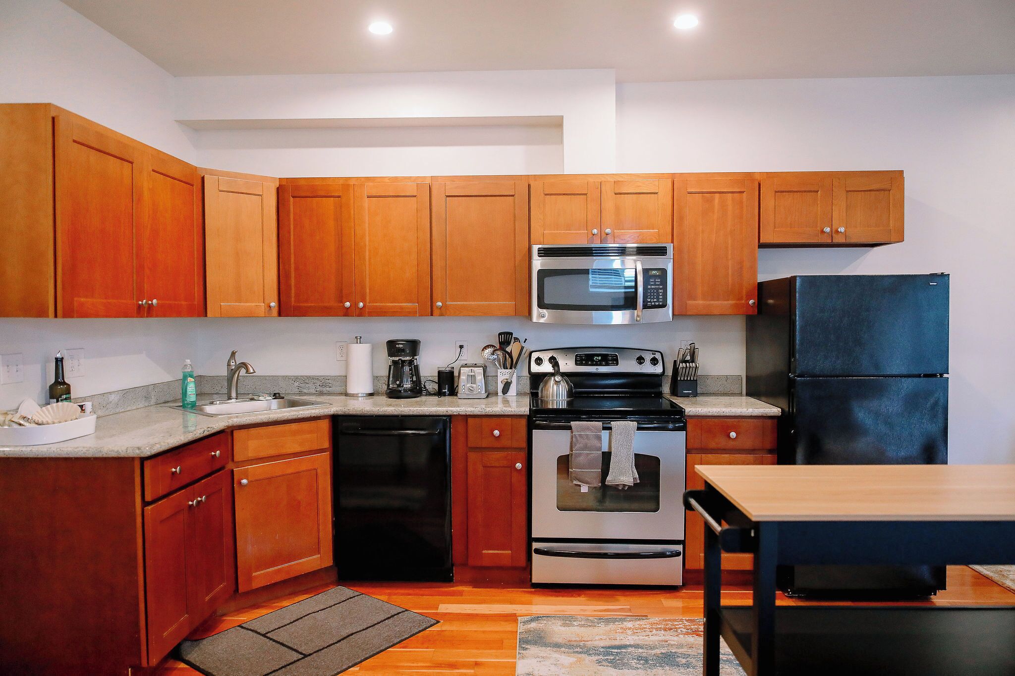 A compact kitchen with wooden cabinets, a stainless steel microwave, a black dishwasher, and a blue refrigerator.