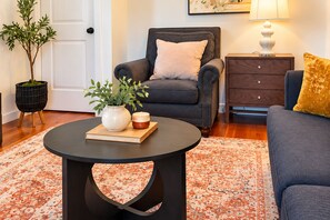 A living room with a dark sofa, a round coffee table with a book and a vase, a side table with a lamp, and a patterned rug.