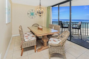 A dining area with a wooden table and wicker chairs, a compass wall decoration, and a view of the beach through large sliding doors.