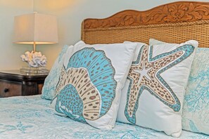 A bed with two decorative pillows featuring sea shell and starfish designs, a wicker headboard, and a bedside table with a lamp and decorative item.