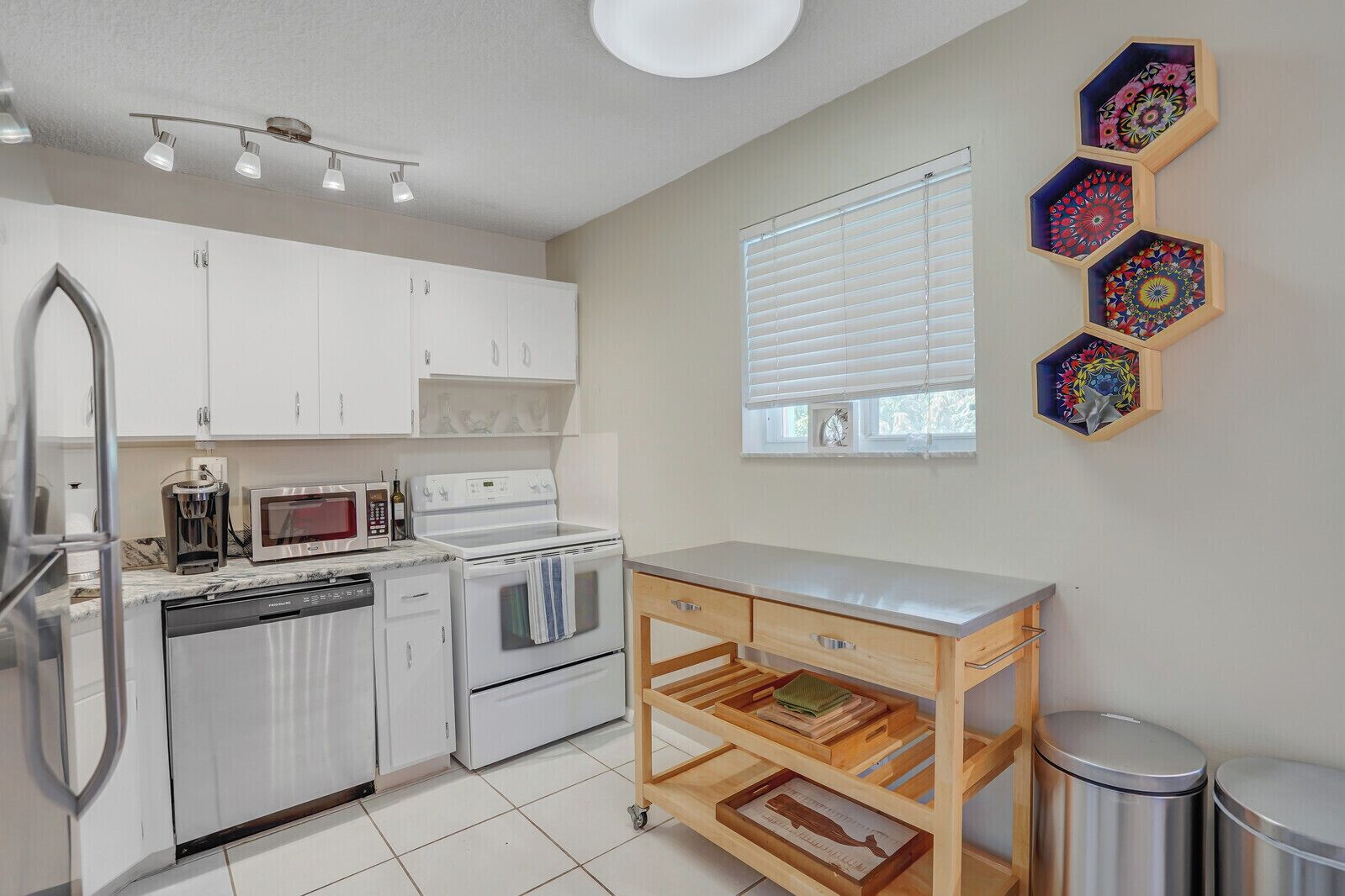 A modern kitchen with white cabinets, a stainless steel refrigerator, a microwave, an oven, and a wooden cart with drawers.