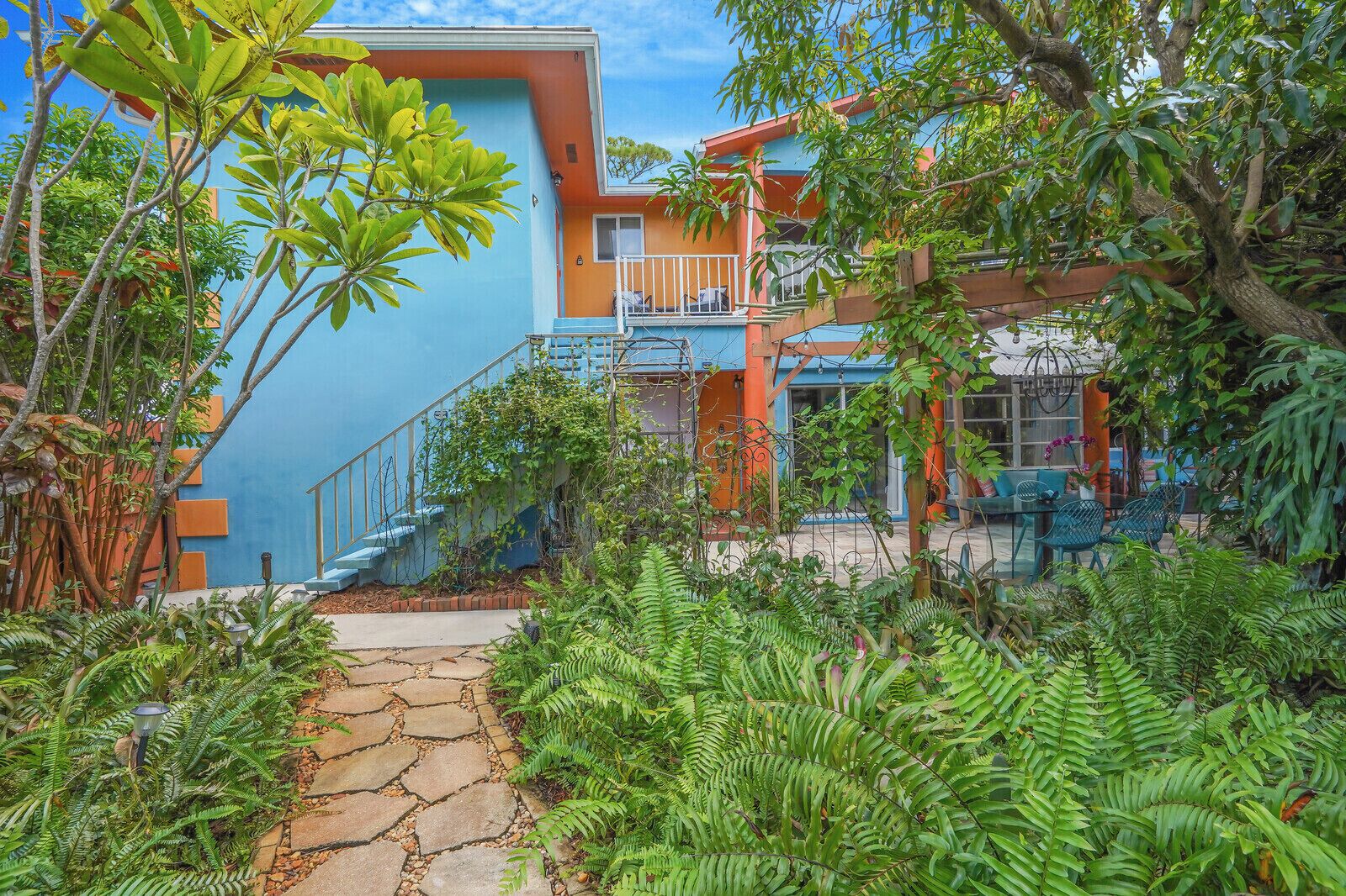 A tropical resort with a stone pathway leading to a blue and orange building, surrounded by lush greenery and a small outdoor seating area.