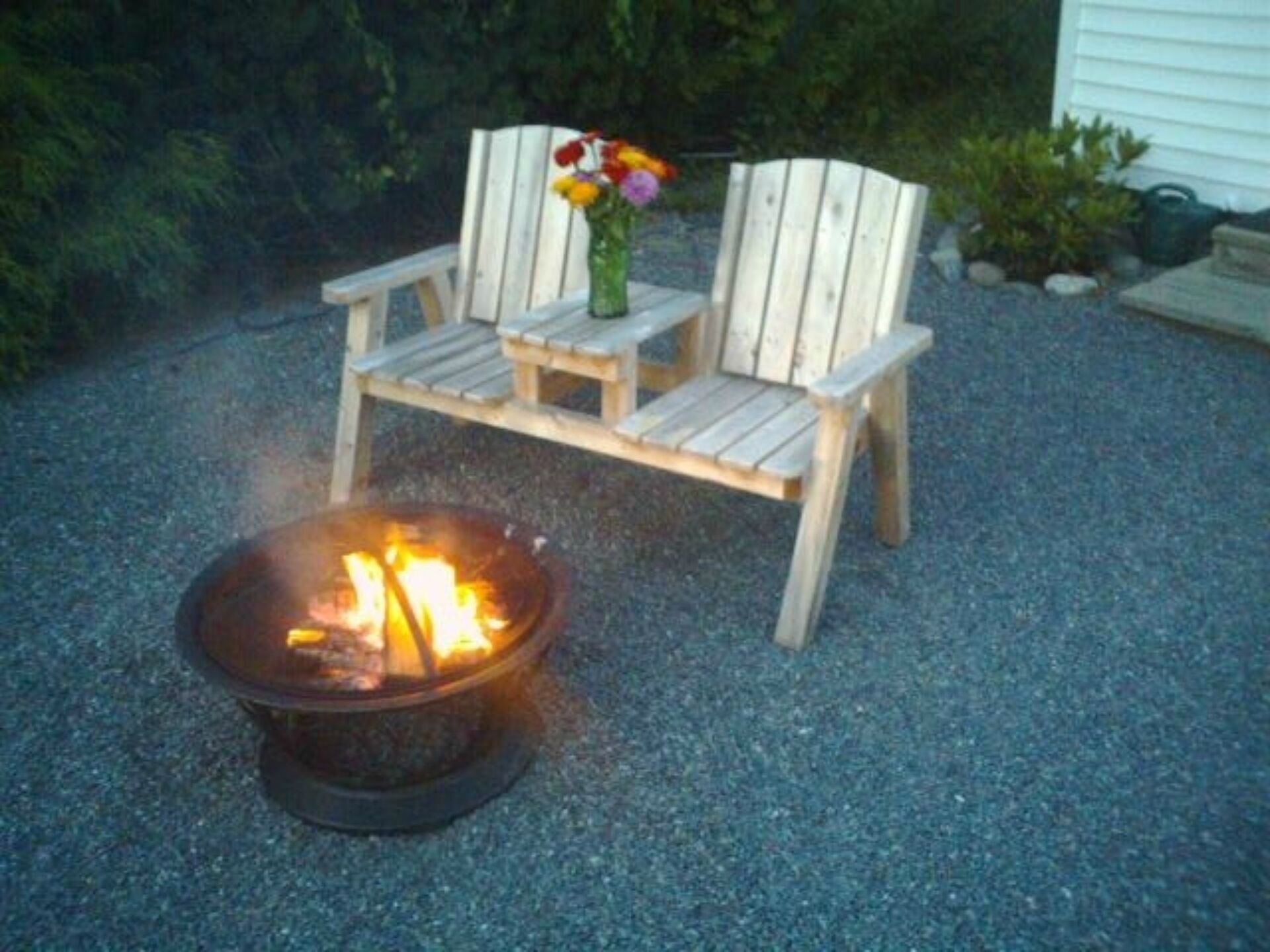 A wooden bench with a vase of flowers, a fire pit, and a gravel ground.
