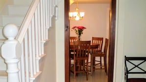 Un escalier avec des balustrades blanches mène à un coin repas où se trouvent des chaises en bois et une table, un lustre et un petit tapis sur le sol.