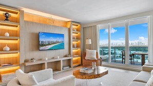 A modern living room with a large TV, comfortable seating, a wooden bookshelf, and a balcony with a city view.