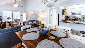 Un salon moderne avec une table à manger en bois, des canapés à motifs bleus et blancs, un ventilateur de plafond et une cuisine dotée de meubles blancs et d’un four à micro-ondes.