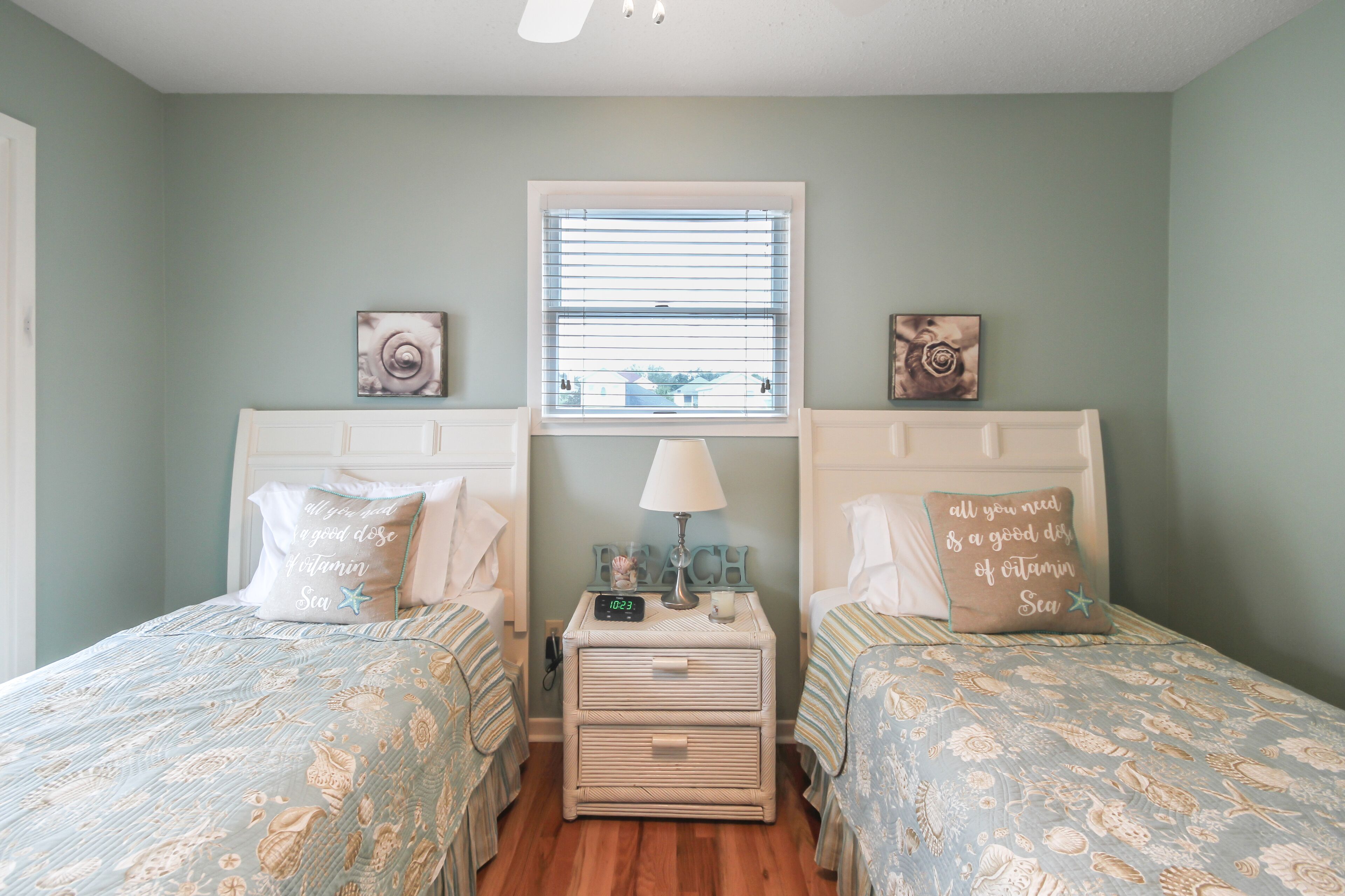 A bedroom with two beds, a nightstand with a clock, a window with blinds, and wall art.