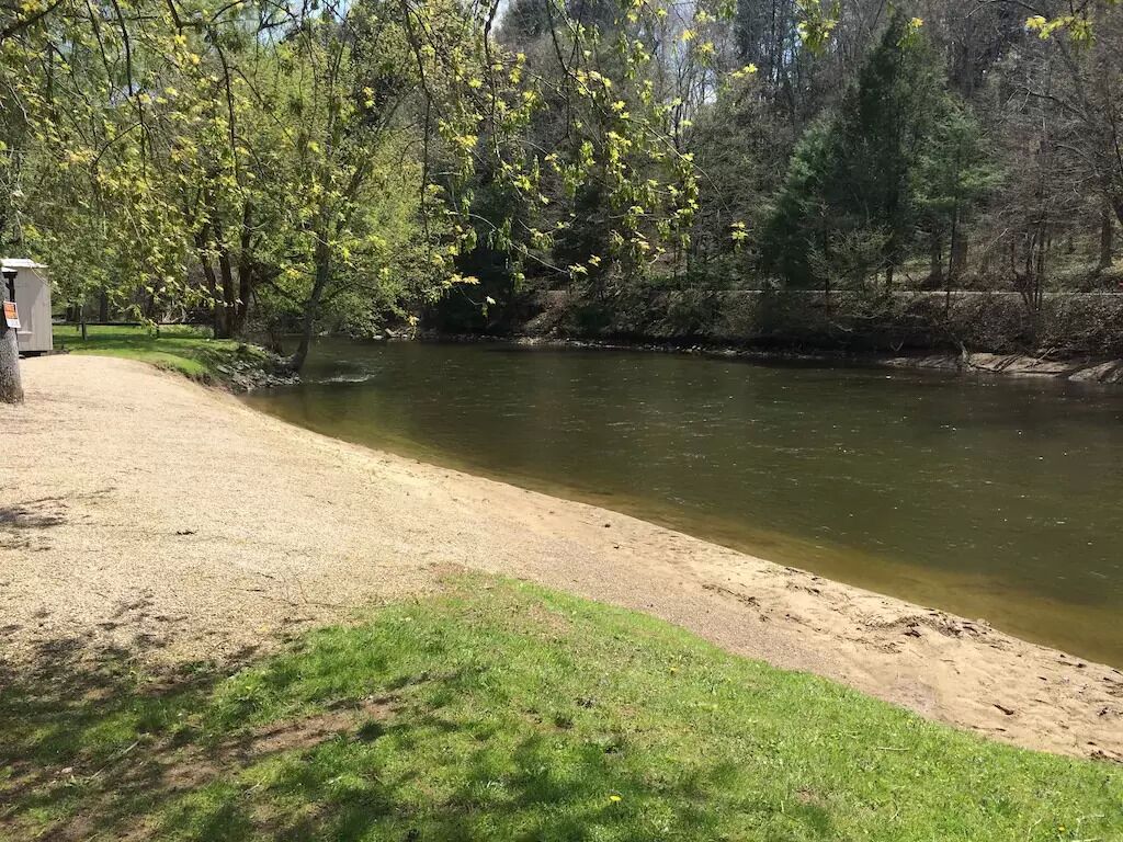 Une rive avec une zone herbeuse et des arbres, une petite construction sur la gauche, et une rivière paisible.