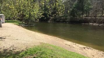 Une rive avec une zone herbeuse et des arbres, une petite construction sur la gauche, et une rivière paisible.
