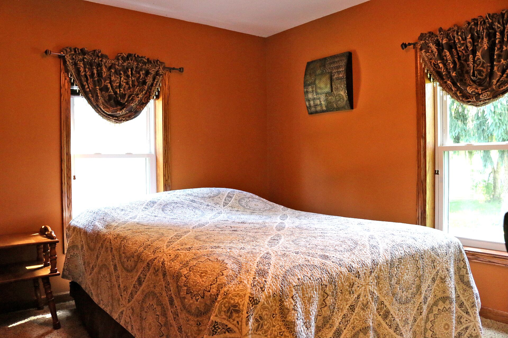 Une chambre à coucher avec un lit, deux fenêtres avec des rideaux, une petite table et un tableau au mur.