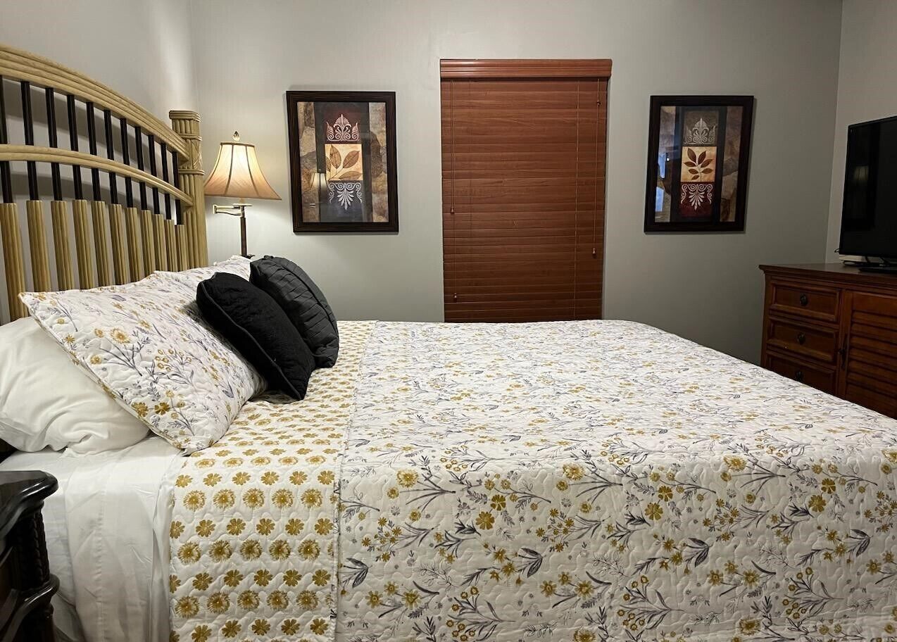 Una cama bien hecha con una colcha floral, un cabecero de madera y una lámpara de noche. Hay dos cuadros en la pared, una puerta de madera y un aparador de madera con un televisor encima.