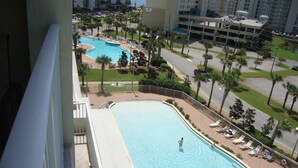 A large outdoor swimming pool with multiple lanes, surrounded by palm trees and lounge chairs, with a view of a parking garage and high-rise buildings in the background.