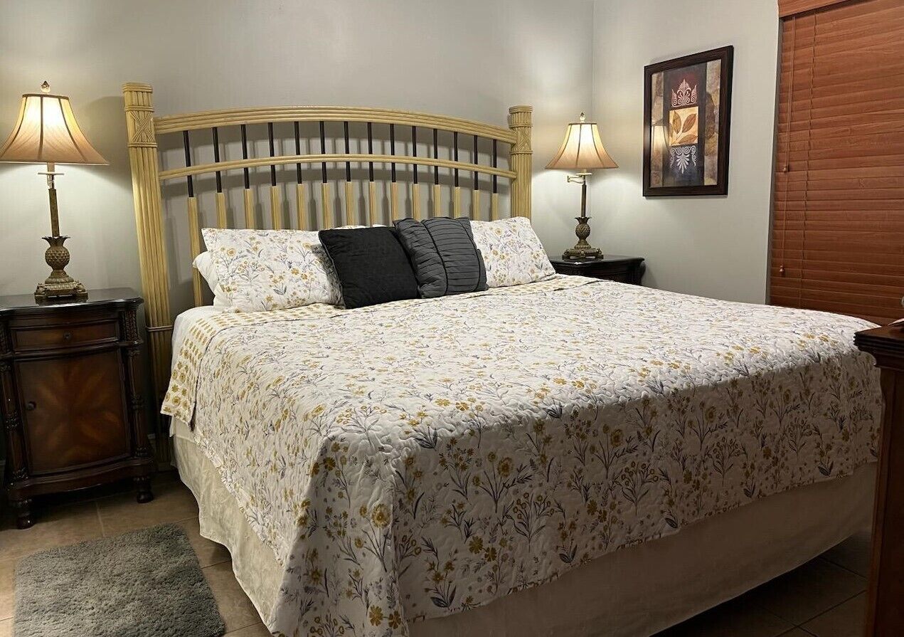 Una cama bien hecha con una colcha estampada, dos lámparas de noche, un cabecero de madera, un cuadro enmarcado en la pared y una puerta de madera.