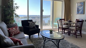 A living room with a sofa, armchair, and a glass-top coffee table. There is a balcony with a view of the water and buildings.