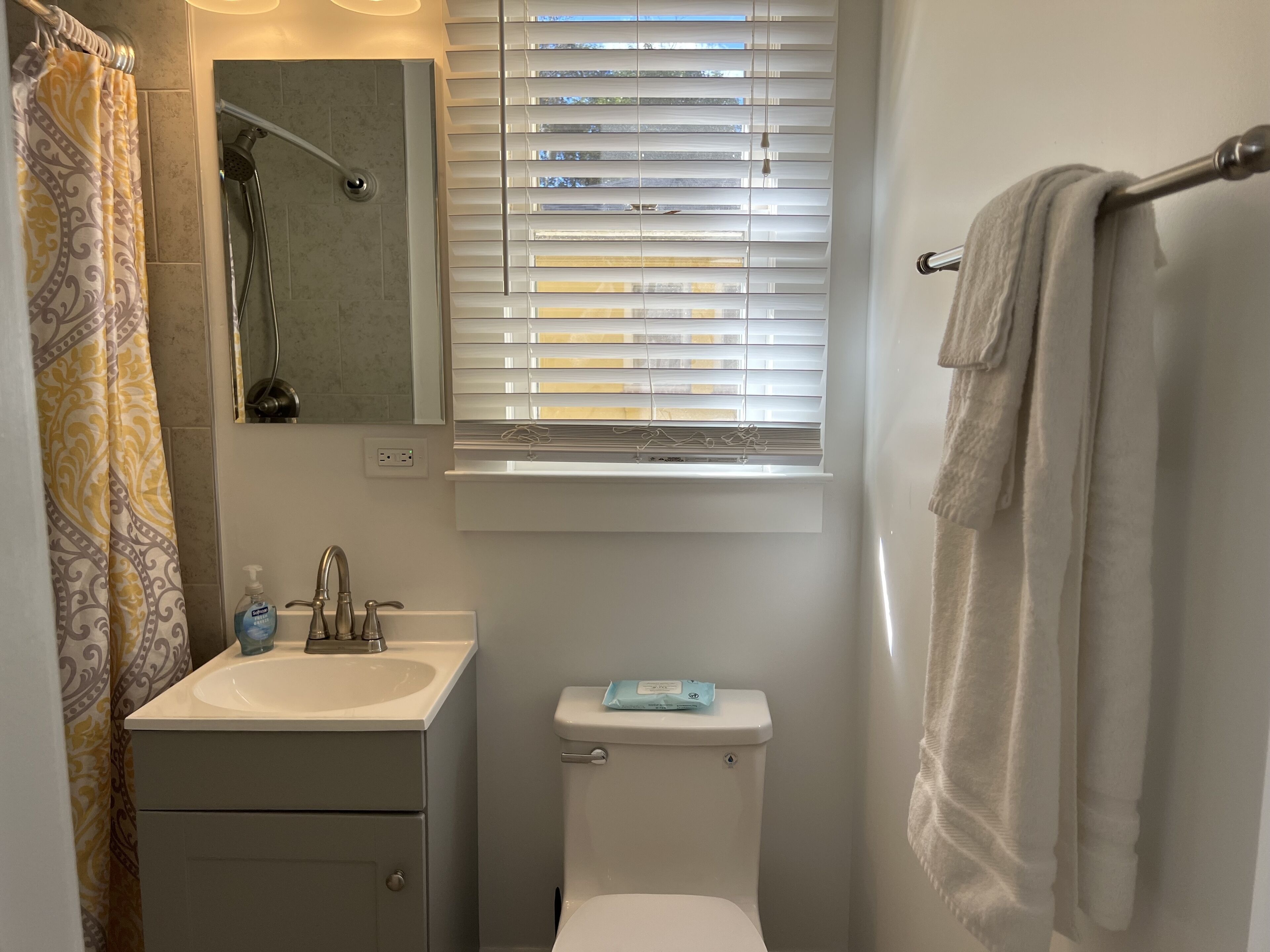 A bathroom with a toilet, sink, shower, and window with blinds.